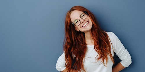 A young female with red hair and glasses tilting her head to the side and smiling